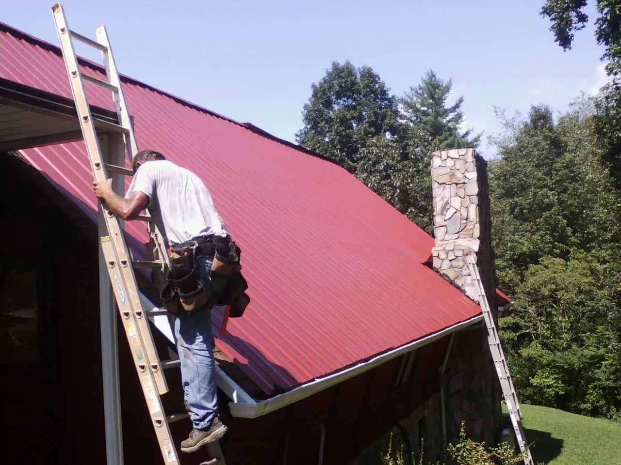 Roofer on a ladder beside a red metal roof and stone chimney — Soffit & Fascia Repair by Roofing Brothers in Avon By The Sea